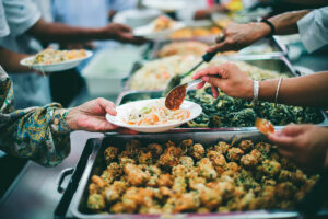 Image showing a buffet line with hands serving food on the right and hands with plates receiving the food on the left. CharityRx Founder's Day of Service