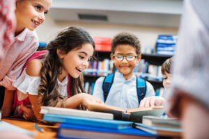 Image of a group of young children reading books in a library. A young girl leans over a book with an interested expression and a young boy in glasses looks on, smiling. CharityRx Founder's Day of Service