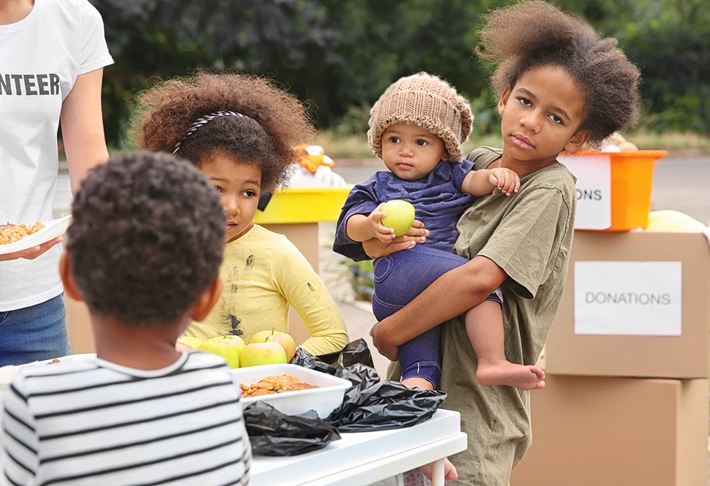 CharityRx helping to reduce food insecurity in America with Feeding Youth USA. Three young Black girls and one Black boy eating from a table staffed by volunteers.