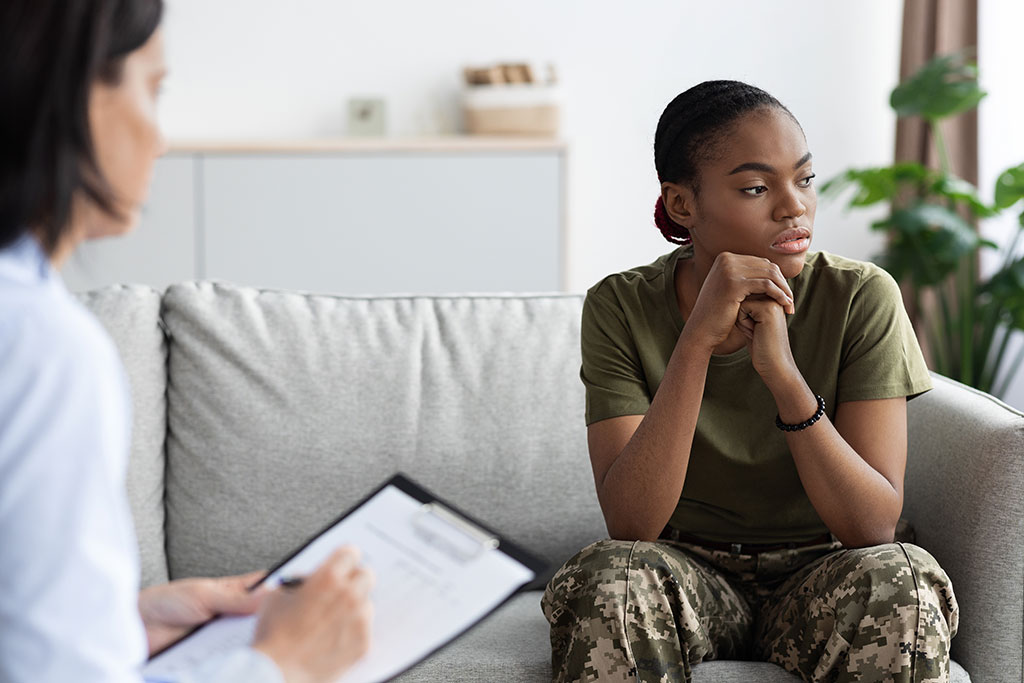 Image of a sad female service member sitting on a couch as a therapist looks on, making notes on a clipboard.