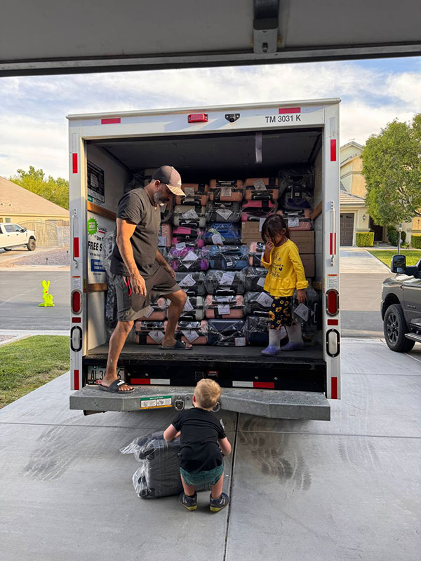 Image of a man and two children loading the back of a U-Haul truck with carry-on sized suitcases CharityRx delivers to Angels of Grace Foster Family Agency.