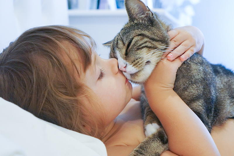 Image of a young girl lying on a bed and kissing the face of a striped cat in her arms.