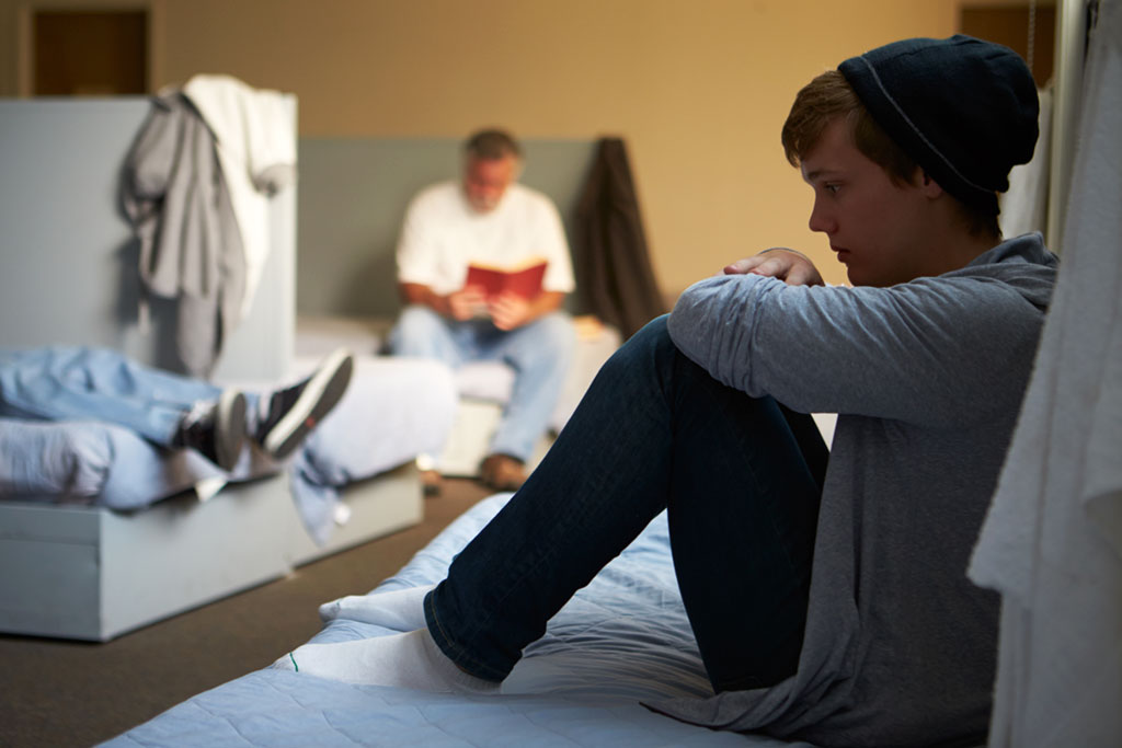 Image of a young teen boy sitting on a bed at a men's homeless shelter, with his knees up to his chest, looking worried. 