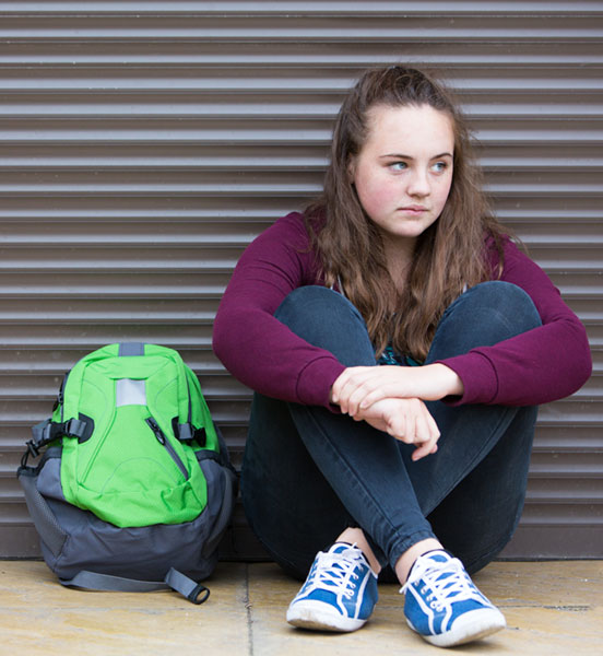 Image of a young teen girl with a sad expression, sitting with knees crossed on the sidewalk in front of a metal grate with a green backpack next to her. 