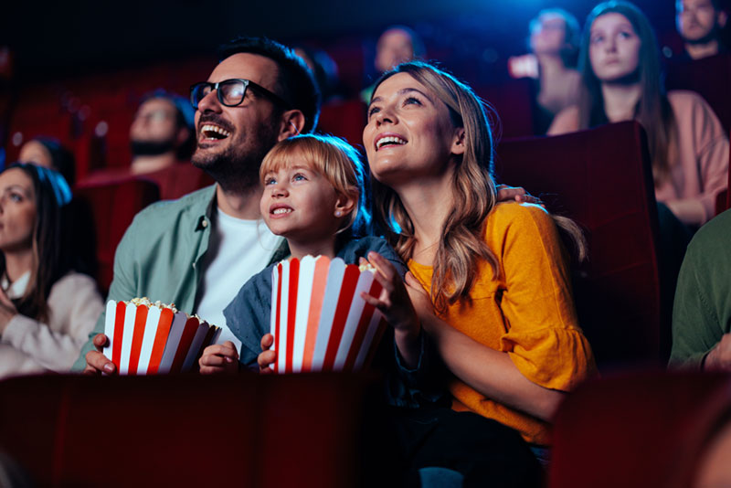 Image of a smiling father, mother, and child in a movie theater, looking up at a screen, holding striped popcorn buckets on their laps.