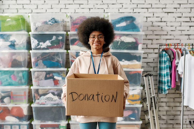 Image of a young smiling black woman in a pink hoodie standing before stacks of plastic bins full of donated clothing. She is holding a cardboard box marked "donation."