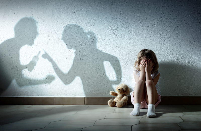 Image of a young girl sitting on the floor next to a teddy bear. Her hands are over her face and next to her, the silhouettes of her fighting mother and father are on the wall. Women with children are especially susceptible to abuse.