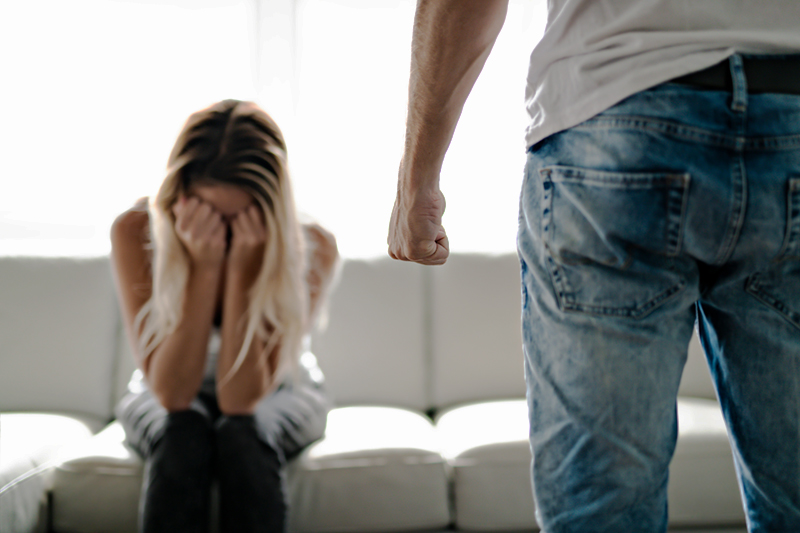 Image of a woman experiencing domestic violence. She is sitting on a sofa with her face in her hands. In the foreground, the lower half of a man stands with one hand in a fist.