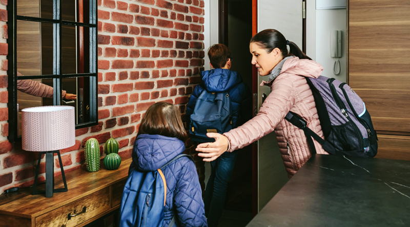 Image of a worried-looking mother shooing her children out the door of their home. All three are wearing coats and backpacks. They are escaping an abusive relationship.