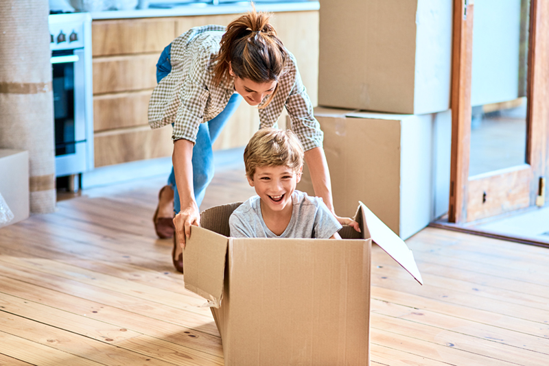 Image of a mother pushing her laughing son across the floor in a cardboard box. They have just moved into a new home after escaping an abusive relationship.