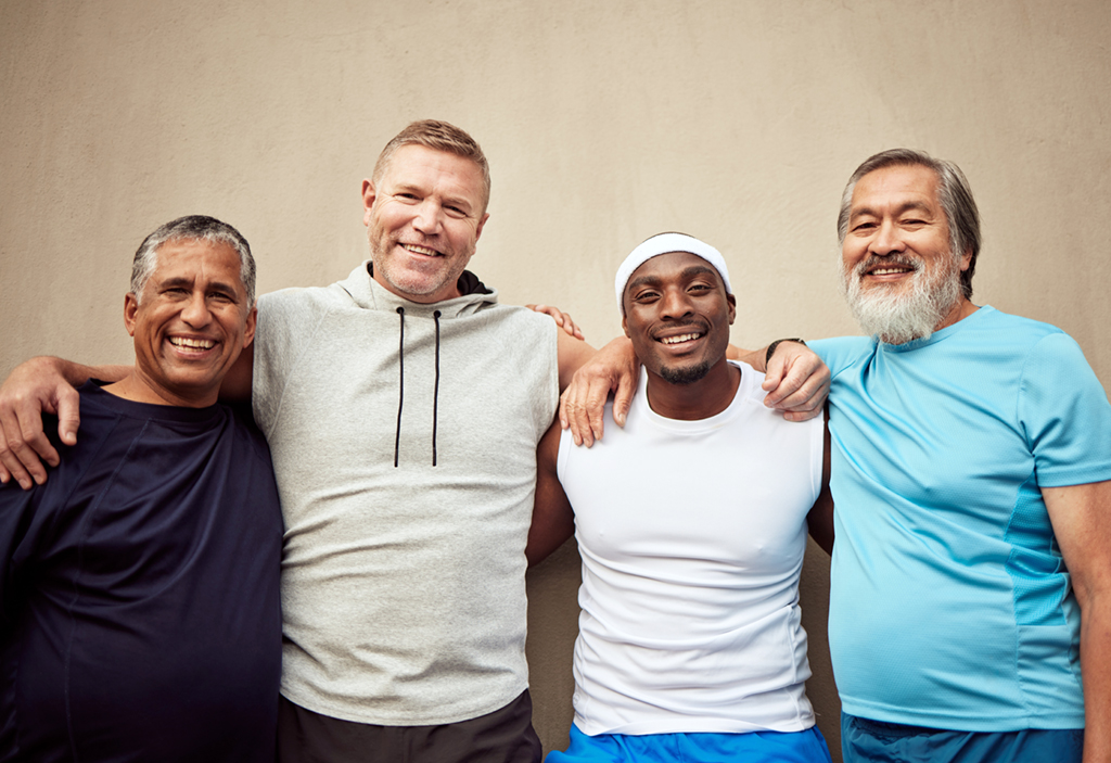 Four smiling men of varying ages and races stand against a wall with their arms over each other's shoulders. 