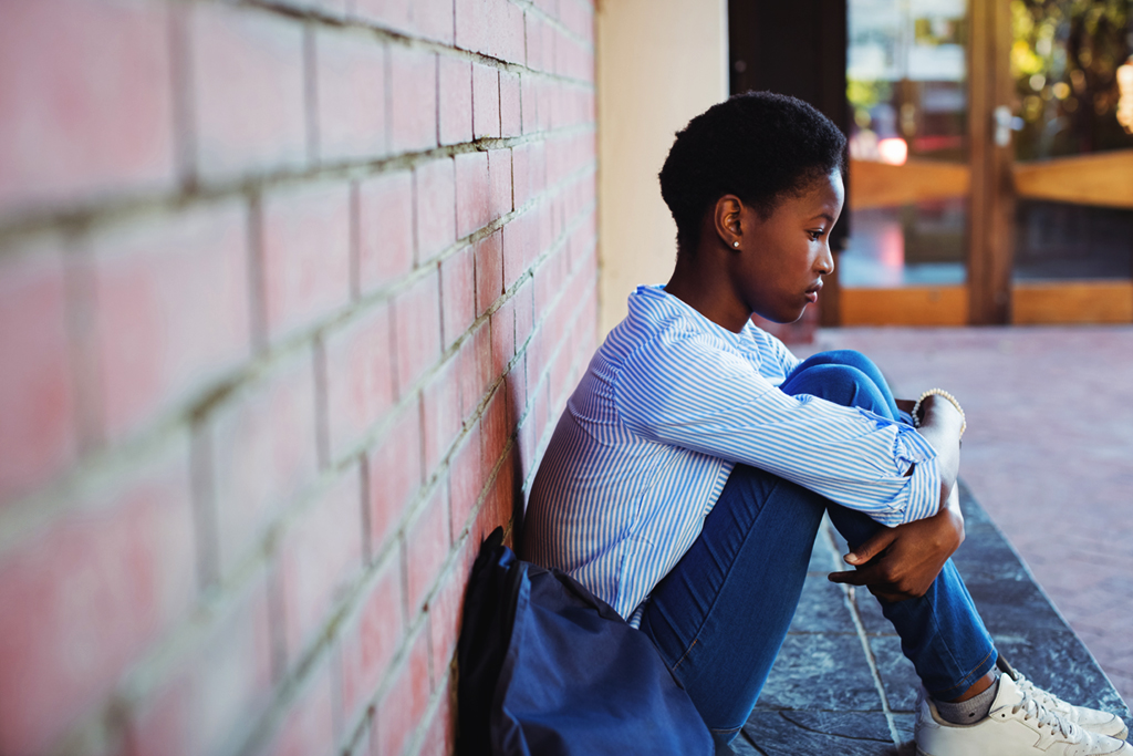 A worried young girl sits alone on a step outside a building, hugging her knees.