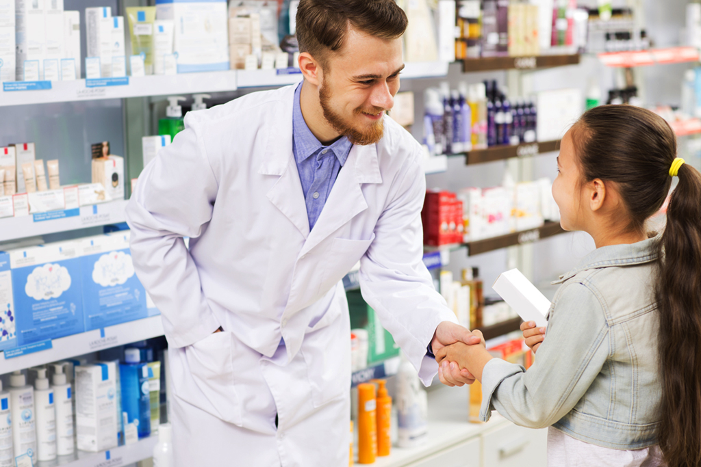 A friendly male pharmacist shakes the hand of a young girl he is helping to find medicine on a shelf.