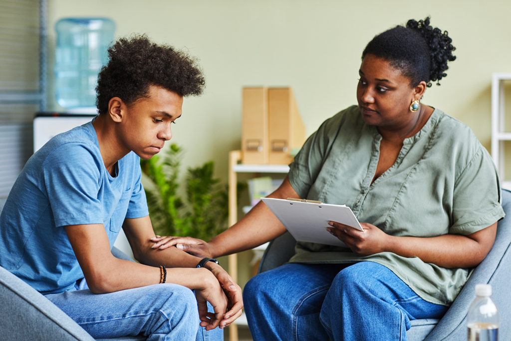 A therapist reaches out and touches the arm of a teen boy who is struggling with his emotions.