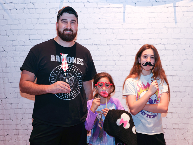 Two young girls and a man hold mustache, lip, and glasses photo booth props and pose for a picture at the movie party event.