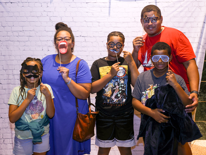 A family of 5 stands together holding photo booth props in front of their faces as they lean together for a photo.