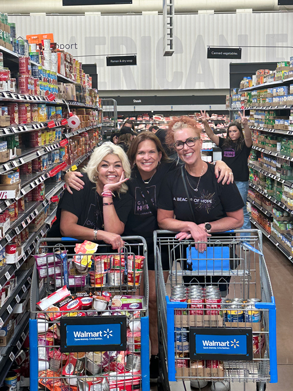 Three smiling women in CharityRx t-shirts lean together behind two shopping carts filled with canned and boxed foods.