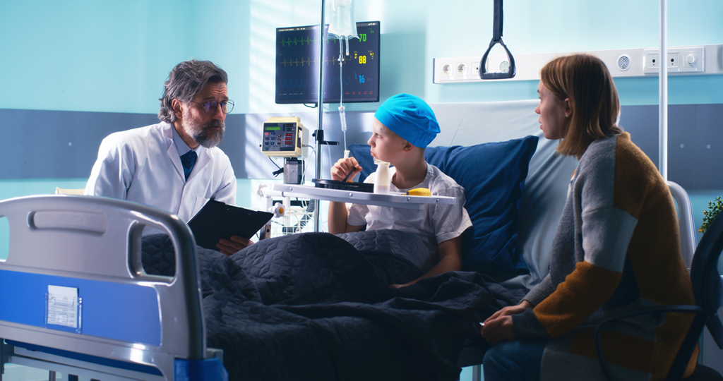 In a hospital room, a doctor speaks to a young boy, who is in the bed wearing a cloth cap to cover his head, and his mother.