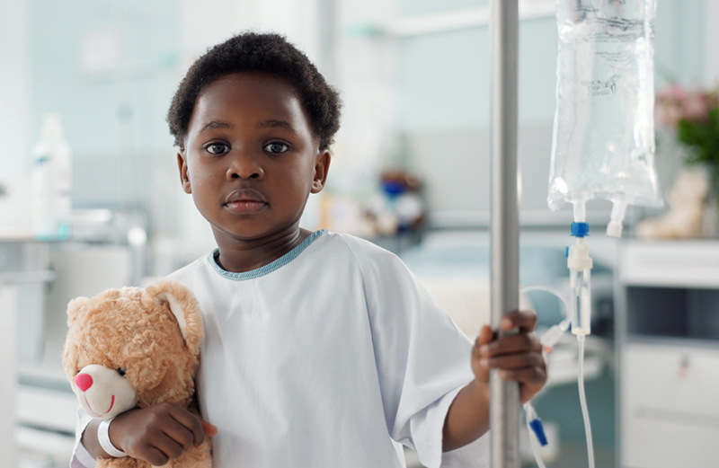 A young boy wearing a hospital gown holds a teddy bear in the crook of one arm and an IV pole in the other hand. He has an IV needle in his hand.