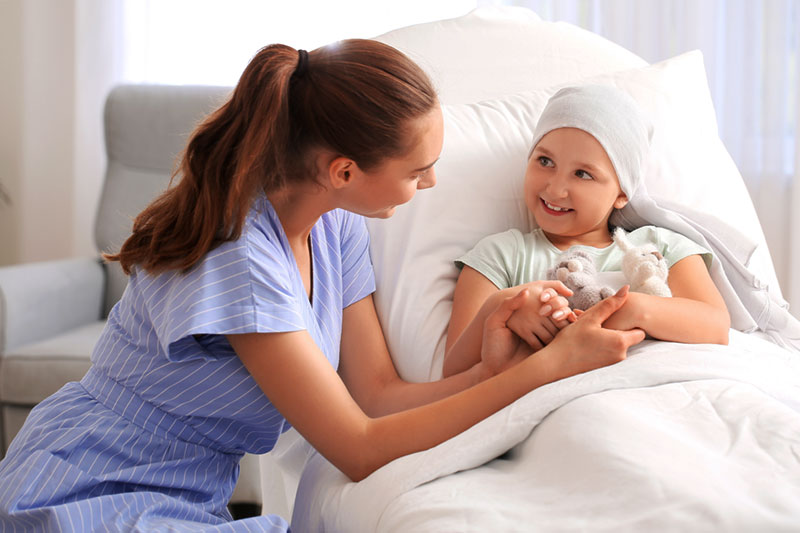 A young girl with cancer in a hospital bed wears a head scarf and holds a stuffed animal. She is smiling as she looks at the nurse who is talking to her and smiling while holding her hands.