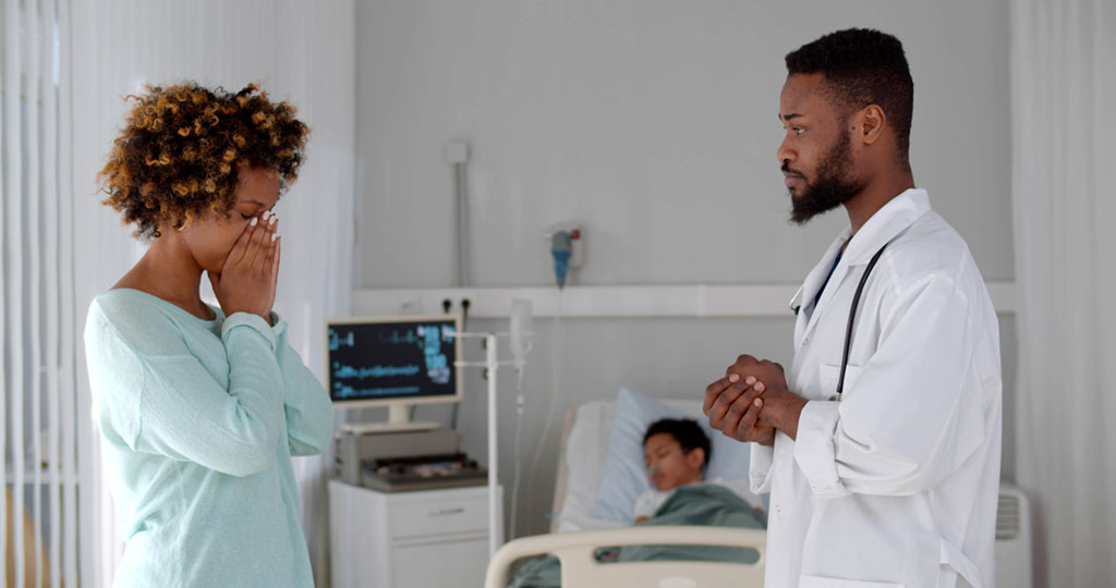 A young mother is distraught at the news her son's physician has delivered as the boy sleeps in a hospital bed in the background. He has monitors and oxygen tubes attached to him.