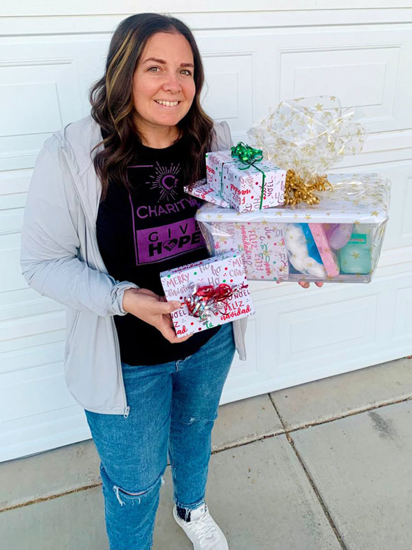 A smiling woman wearing a CharityRx t-shirt holds several wrapped gifts and a bin of nail care supplies.