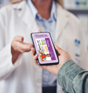 A hand holding a smart phone with a CharityRx prescription discount card on the screen shows the image to a pharmacist.