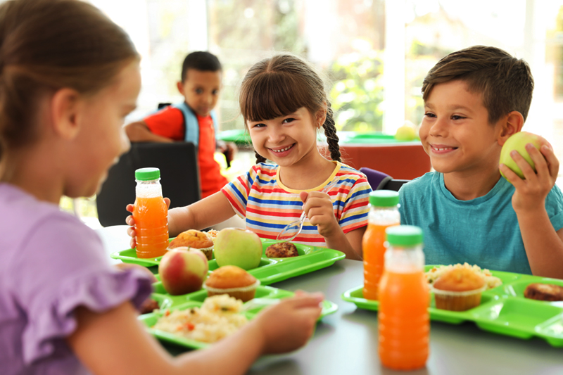 Happy young children gather around a lunch table at school. Each child has a tray full of food in front of them. 