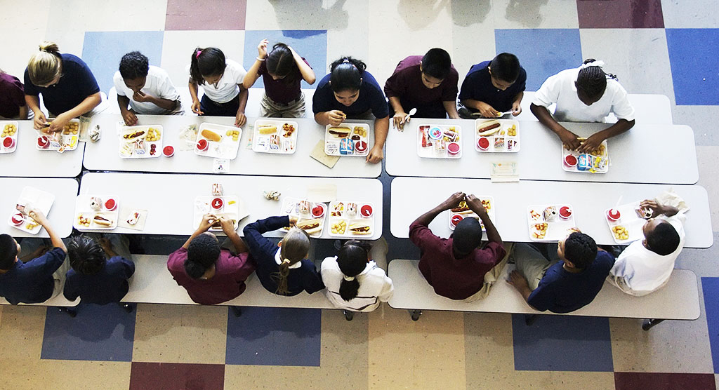 Aerial view of a school lunch table with about 20 children eating lunch from trays of food.