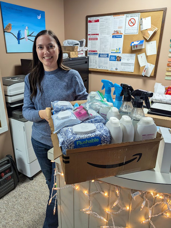 A young woman smiles as she holds a box full of hygiene and cleaning supplies donated to Sarah's Friends.