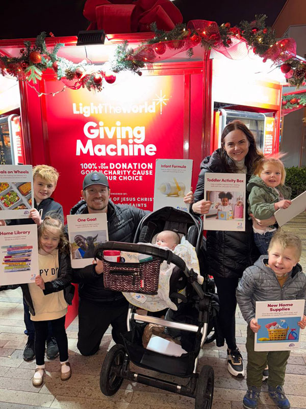 A young family with five young children stands in front of a Light the World Giving Machine holding cards for the items they donated.