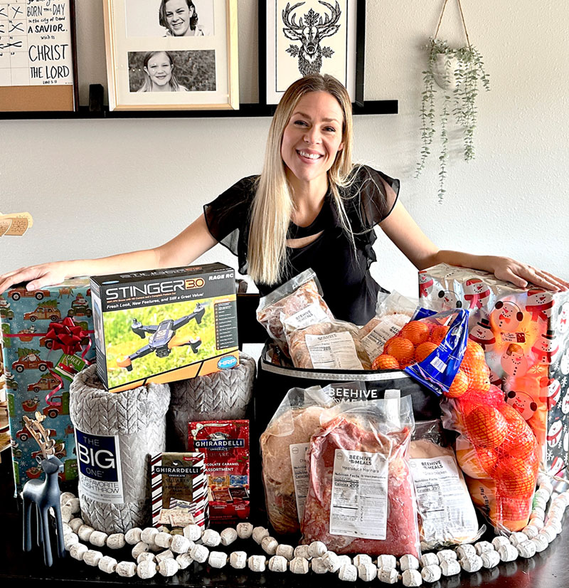 A smiling young woman stands behind the food and gifts she purchased to donate to families in her neighborhood.