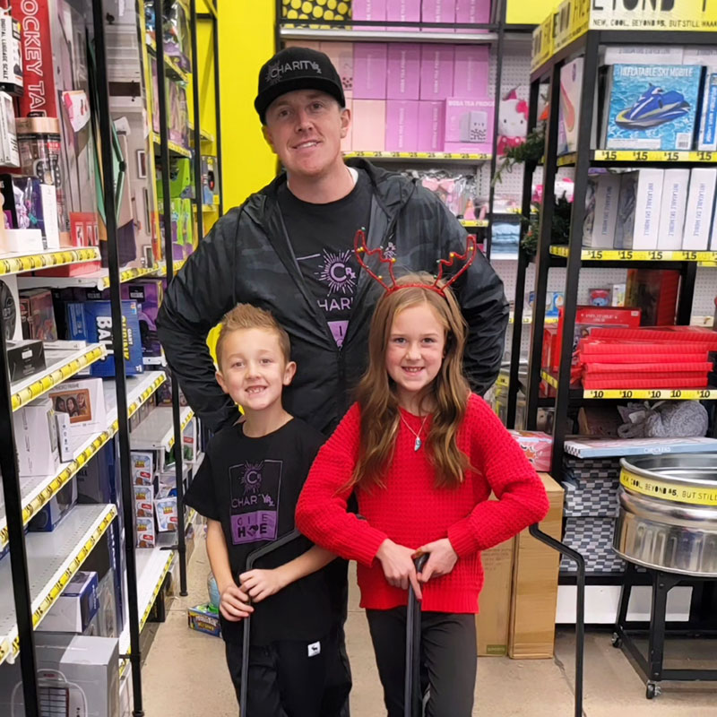 A father stands behind his son and daughter while shopping for gifts to donate.