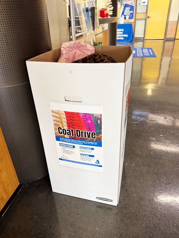 A large box of winter coats in the hallway of a school with a label reading "Coat Drive"