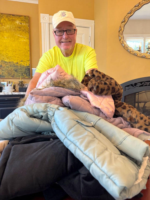 A man wearing a CharityRx hat stands behind a large pile of winter coats of different colors and styles.