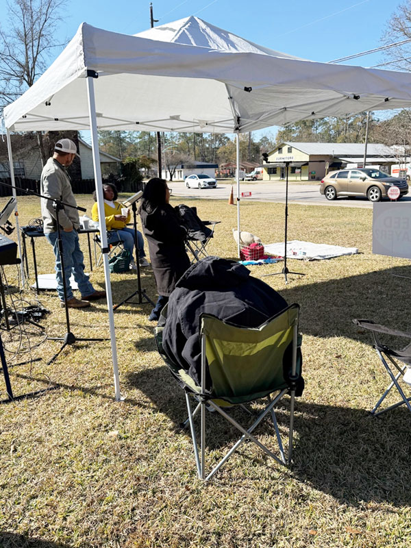 A man and two women set up microphones and speakers under a tent in a local park.